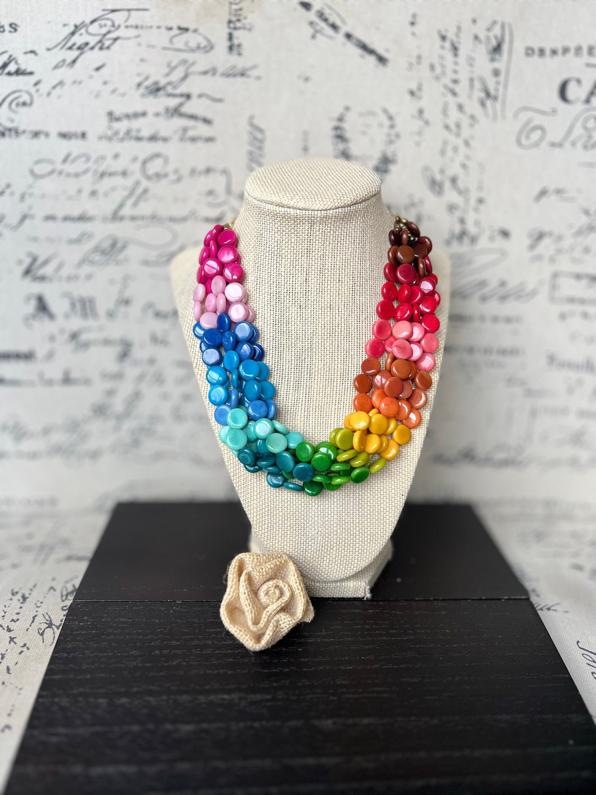 May include: A rainbow colored necklace made of wooden beads. The necklace is displayed on a white mannequin. A small crocheted flower is in the foreground.