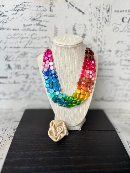 May include: A rainbow colored necklace made of wooden beads. The necklace is displayed on a white mannequin. A small crocheted flower is in the foreground.