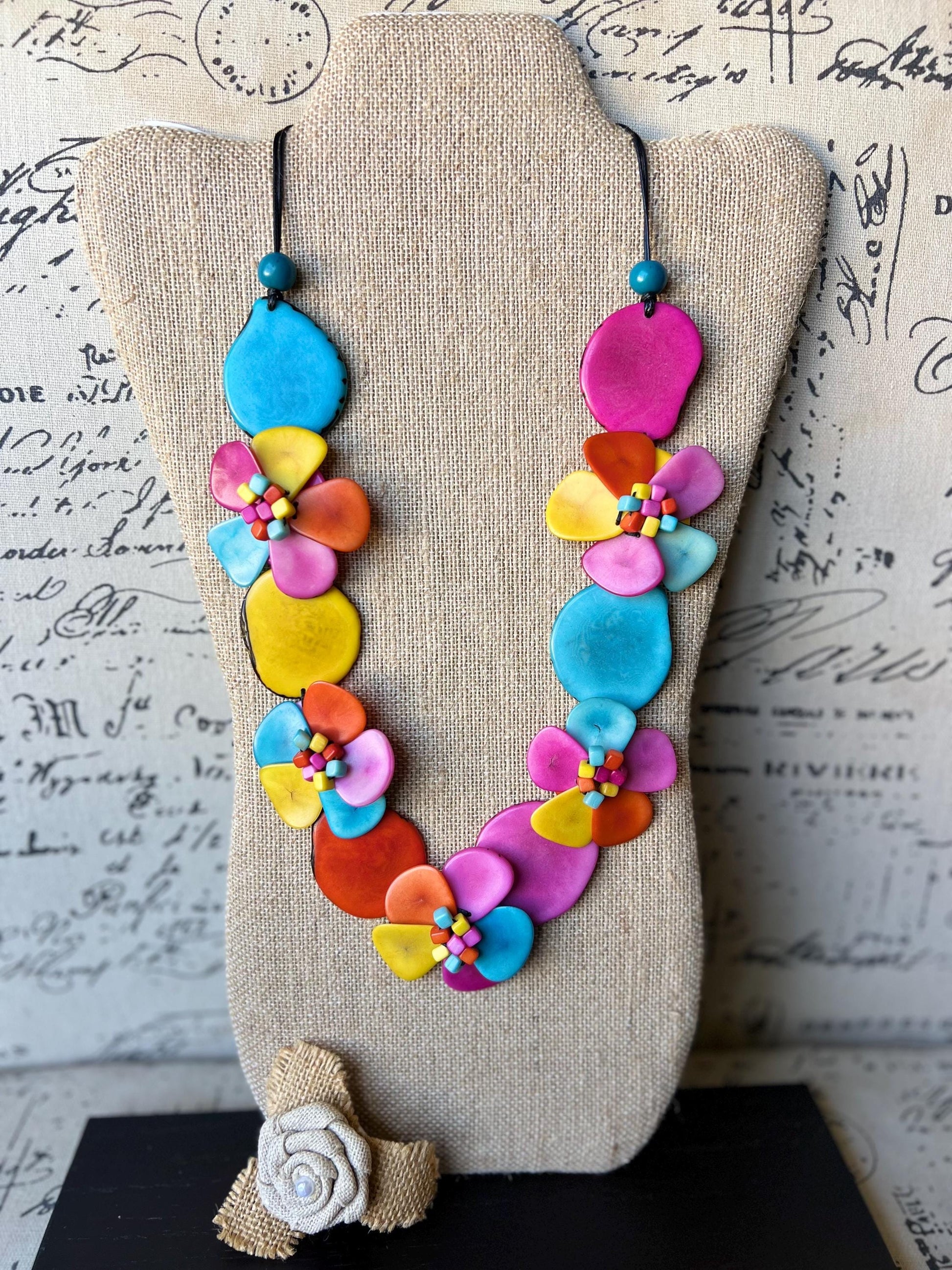 May include: A colorful necklace made of layered, painted, flower-shaped ceramic pieces. The necklace is displayed on a beige fabric-covered mannequin. The necklace features shades of pink, yellow, blue, orange, and red.