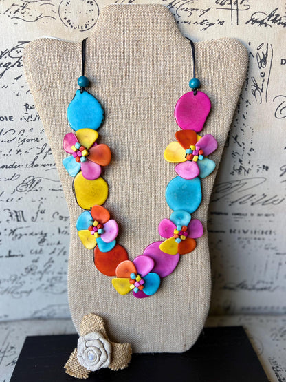 May include: A colorful necklace made of layered, painted, flower-shaped ceramic pieces. The necklace is displayed on a beige fabric-covered mannequin. The necklace features shades of pink, yellow, blue, orange, and red.