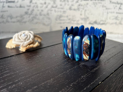 May include: A blue and white bracelet made of oval shaped beads. The bracelet is made of a dark blue material with white and brown swirls. The bracelet is on a dark wooden surface.